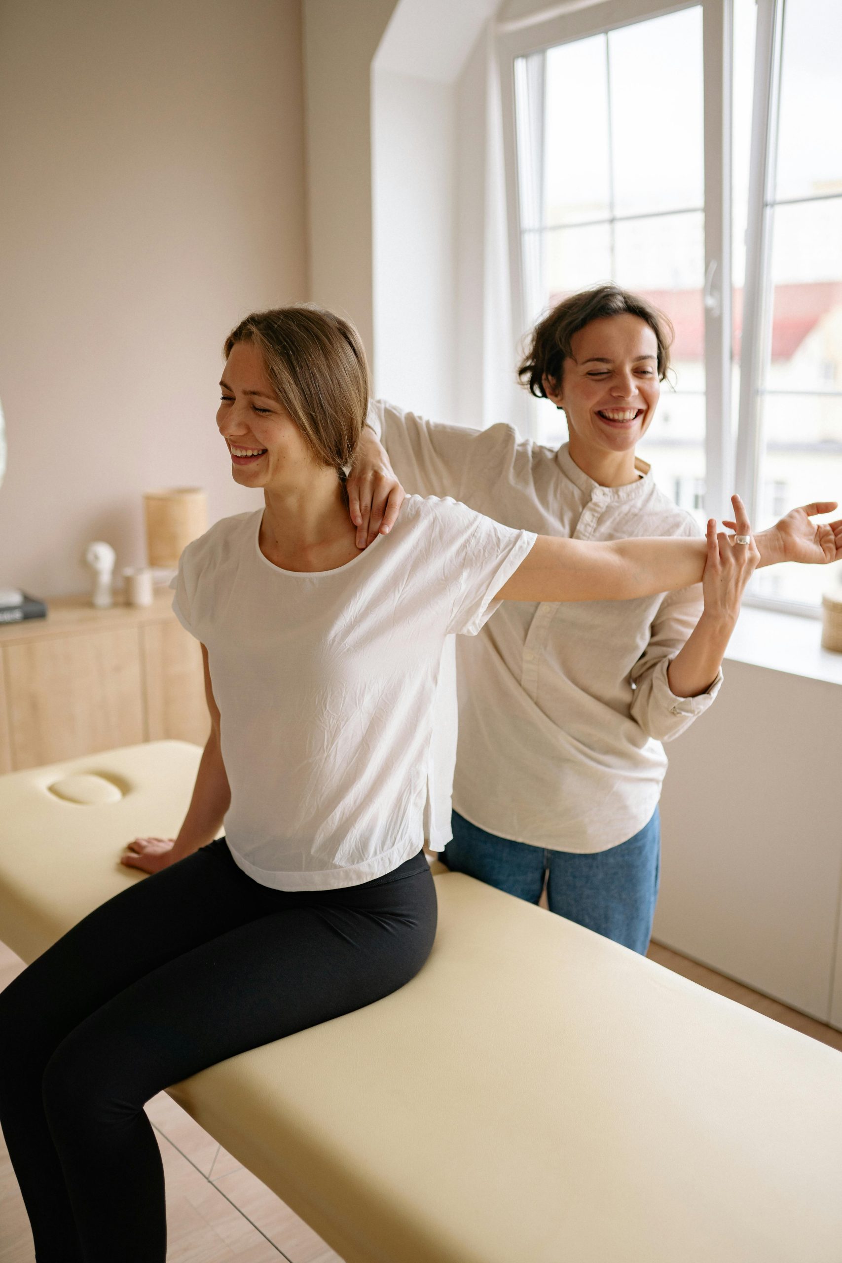 A therapist guides a woman in a stretching exercise on a massage table in a clinic setting.